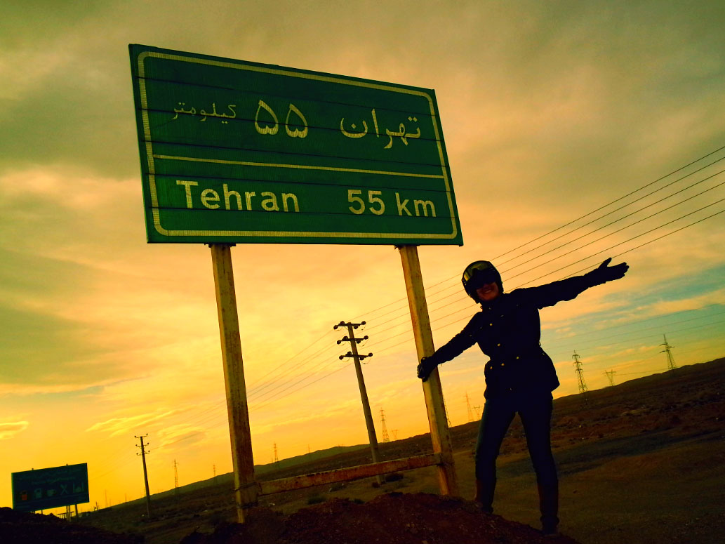 After dinner speaker Lois Pryce with her arms outstretched next to a road sign saying she is 55km outside of Tehran, Iran